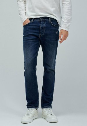 Jeans de mezclilla azul oscuro con un corte recto clásico, que presentan un ligero desgaste y desgarros sutiles en la parte delantera, combinados con zapatillas blancas.