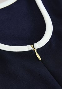 Nahaufnahme eines marineblauen Stoffes mit weißem Saum am Ausschnitt, der einen kleinen goldenen Reißverschluss-Schieber teilweise geschlossen zeigt.