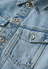 Veste en denim bleu clair avec poches poitrine boutonnées et boutons argentés visibles sur tissu texturé.