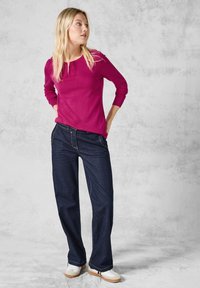 Maglione a maglia fucsia con maniche lunghe, abbinato a jeans a gamba larga blu scuro. Footwear composto da sneaker bianche con accenti di suola color gomma. Texture lisce.