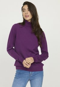 Chandail col roulé violet en tissu doux, avec des manches longues et des bords côtelés. Associé à un jean bleu, sur fond clair.