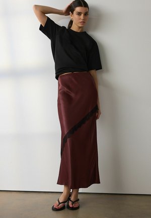 Naine kannab musta üle suurusega t-särki, burgundia satiinist seelikut musta pitsiga ja mustasid sandaale, seisab valge seina ees.