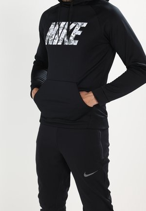 Person trägt schwarzen Nike-Hoodie mit Tarnmuster-Logo und passende schwarze Nike-Hose mit Swoosh-Logo, Hände in der Kapuzenbeuteltasche.