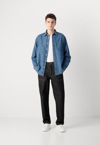 Camicetta di denim blu con chiusura a bottoni e tasca, abbinata a pantaloni neri a vita alta e sneakers bianche, su uno sfondo semplice.