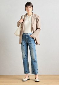 Beige blazer over en hvit ribbet polo, blå jeans med blå blomsterdetaljer og kremfargede loafers. Modell som holder en lys skulderveske.