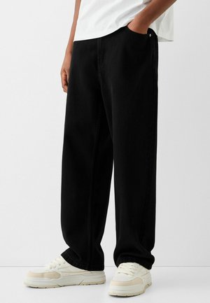 Personne portant un pantalon noir ample et des baskets blanches, debout avec une main dans la poche devant un fond uni.