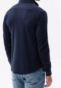 Marinblå långärmad skjorta med knäppkrage, slät textur och subtila detaljer på baksidan och ärmarna, ihop med ljusblå jeans.