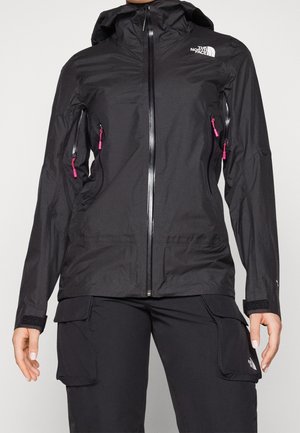 Persoon draagt een zwarte waterdichte The North Face jas met capuchon en ritszakken, gecombineerd met zwarte cargobroek met klepzakken.