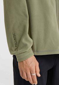 Green fleece jacket with Polartec branding, featuring a ribbed cuff and a soft texture. Visible stitching detail along the hem.