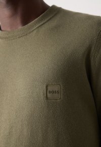 Maglione a maglia verde oliva con scollo rotondo e tessuto texturizzato. Toppa con logo quadrato ricamato sul petto.