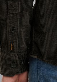 Chaqueta de pana marrón oscuro con una textura acanalada, que cuenta con un cierre de botón y una etiqueta con emblema. Los puños tienen dos botones.