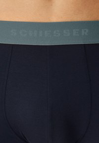 Granatowe slipy męskie wykonane z gładkiego materiału, z miętowozieloną elastyczną talią z logo "SCHIESSER" w kontrastowej fakturze.
