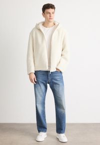 Bunda s kapuljačom od flisa u krem boji, s patentnim zatvaračem sprijeda i dugim rukavima. Uparena s svijetloplavim trapericama ravnog kroja i bijelim cipelama.