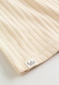 Tissu beige côtelé avec une petite étiquette blanche cousue portant un logo de marque en cursive noire sur un fond clair.