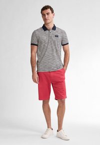 Pruhované polo tričko v bílé a námořnické modři s límečkem, spárované s červenými šortkami. Model má na sobě bílé tenisky. Jednoduchý, casual a funkční outfit.