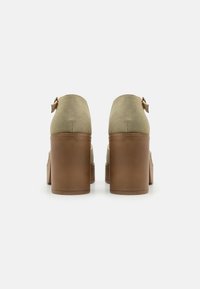 Scarpe alte in suede verde con un ampio tacco in legno marrone e un sottile cinturino per la caviglia con dettaglio della fibbia dorata.