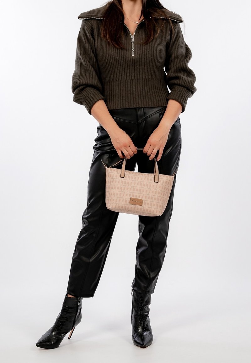 Femme portant un pull côtelé foncé et un pantalon en cuir noir, tenant un sac à main beige de créateur et portant des bottines noires à talons hauts.