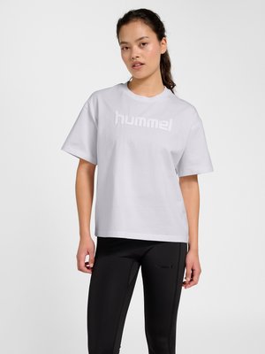 Kvinde med mørkt hår i hestehale iført en hvid løs Hummel t-shirt og sorte leggings mod en ensfarvet lys baggrund.