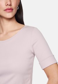 Junge Frau trägt ein helllila kurzärmliges Shirt mit Rundhalsausschnitt und silbernen Creolen, von den Schultern bis zu den Lippen gezeigt.