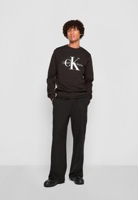 Svart hettegenser med en hvit "Calvin Klein Jeans" logo og "CK" grafikk. Sammen med svarte, avslappede cargo-bukser og kraftige sko.