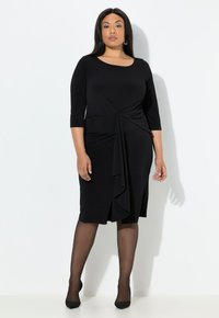 Vestido negro a la rodilla con mangas tres cuartos, que presenta un detalle drapeado en la parte delantera y una textura suave. Combinado con medias negras transparentes y tacones.