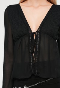 Zwarte blouse met lange mouwen, diepe V-hals, kanttextuur op het bovenste deel en striksluiting. Doorzichtige stof met delicate afwerkdetails.