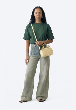 Femeie purtând un tricou verde, blugi evazați decolorati, sandale negre, cercei aurii cerc și o geantă bej de tip crossbody cu mânere.