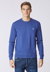 Maglione blu a collo rotondo realizzato in tessuto morbido, con maniche lunghe e un piccolo logo sul petto. Indossato con jeans blu, su uno sfondo neutro.