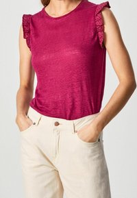 Žena v bezramínkovém malinovém topu s volánky na ramenou a v kalhotách s vysokým pasem v barvě špinavé bílé, s rukama v kapsách.