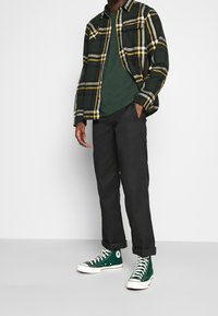 Uomo che indossa una camicia a quadri verde scuro e gialla, una maglietta verde scuro, pantaloni neri e sneakers alte verde scuro con lacci bianchi.
