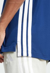 Blauwe sporttrui met een witte drie-strepen ontwerp langs de zijkant. Gemaakt van gestructureerde stof, met zijsplitten voor bewegingsgemak.