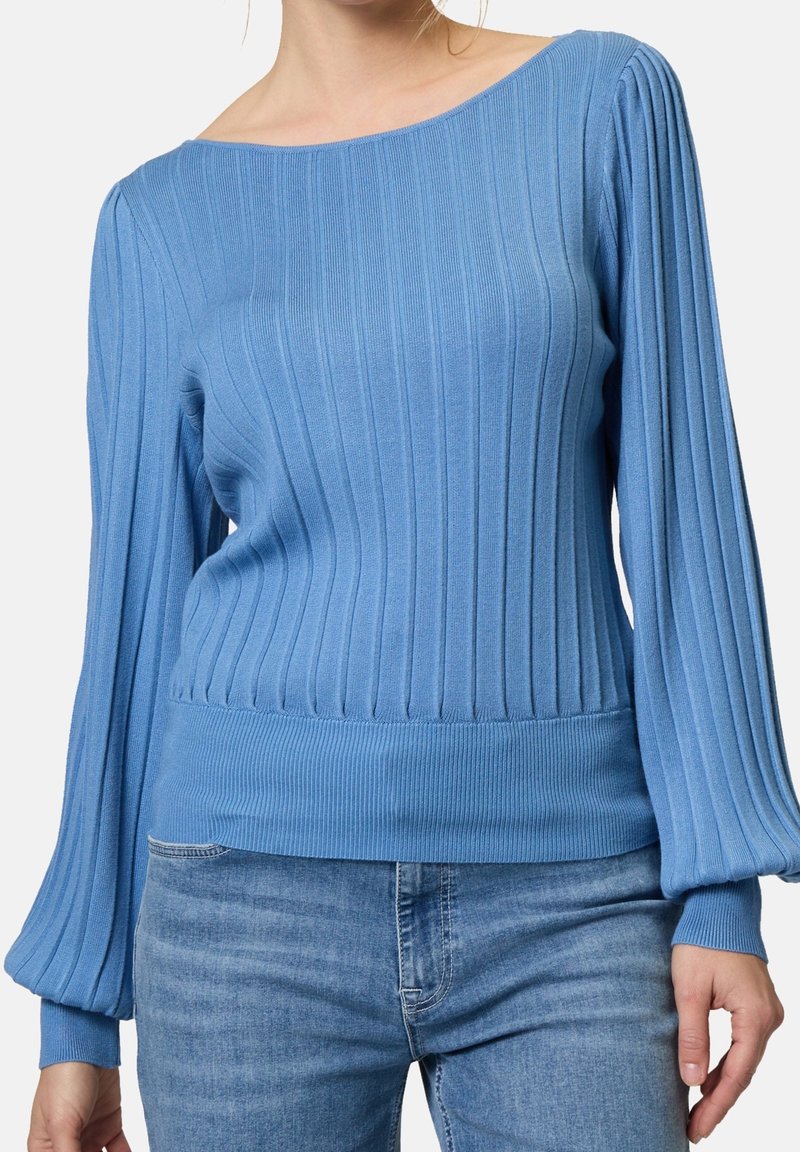 Femme portant un pull en maille côtelée bleu clair avec des manches ballon longues, associé à un jean denim de lavage moyen, debout devant un fond uni.