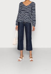 Donna che indossa una blusa a maniche lunghe navy e bianca con un motivo, jeans larghi cropped scuri, sandali con tacco bianchi e tiene in mano una piccola borsa bianca.