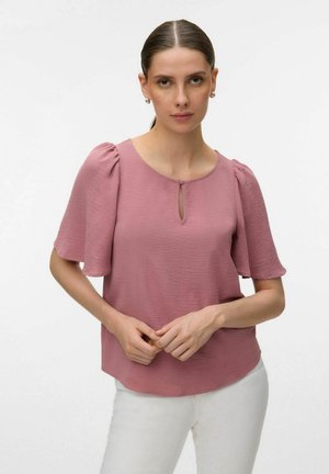 Femme aux cheveux lissés en arrière, portant un chemisier ample rose poussiéreux à manches volantées et un pantalon blanc, debout devant un fond uni.