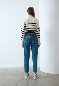 Krämfärgad tröja med svarta ränder, hög krage; matchad med lösa blå denimjeans och sportskor med metalliska detaljer.