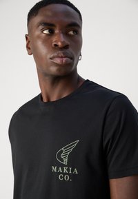 Black cotton t-shirt featuring a green wing logo and "MAKIA CO." text on the front. Classic crew neck design, soft texture.
