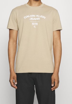 T-shirt beige à manches courtes avec le texte blanc "Calvin Klein Jeans 1978" porté par une personne avec un pantalon noir sur un fond uni.