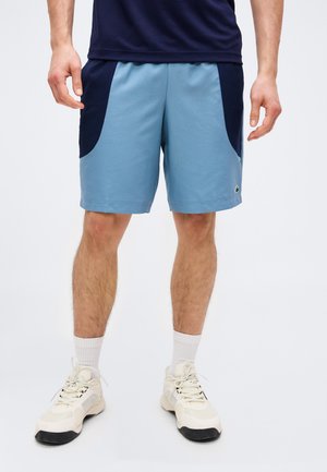 Man die lichtblauwe en marineblauwe korte broek, witte sokken en witte sneakers draagt, staand tegen een effen achtergrond.