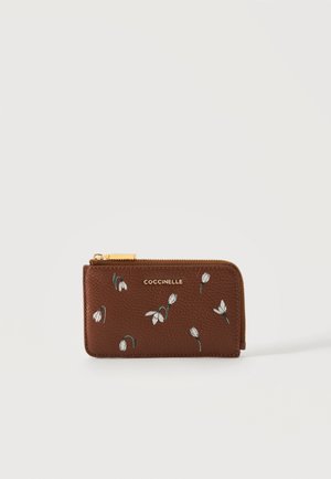 Braune Leder-Geldbörse mit Reißverschluss, kleiner weißer Blumenstickerei und goldenem "COCCINELLE"-Logo vorne vor einfarbigem Hintergrund.