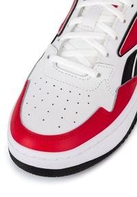 Detailný záber na bielu tenisku s červenými a čiernymi akcentmi, perforovanou špičkou a bielymi šnúrkami na textúrovanej čiernej podrážke.