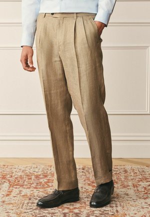 Man wearing beige pleated trousers, light blue shirt, and black woven leather loafers, standing on patterned rug against paneled wall.