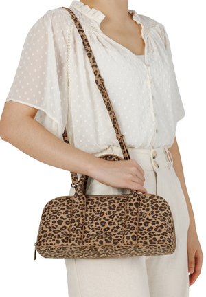 Frau trägt weiße Bluse und Hose und hält eine rechteckige Handtasche mit Leopardenmuster, Henkel oben und Schulterriemen.