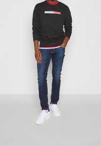 Man som bär en mörk långärmad Tommy Jeans sweatshirt, blå jeans och vita sneakers, står med ena handen i fickan.