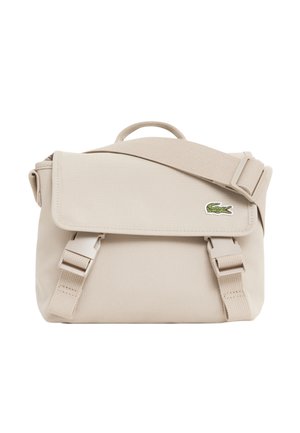 Sac à bandoulière en tissu beige avec rabat, deux sangles avant avec boucle, une poignée supérieure et un petit logo crocodile vert sur le rabat.
