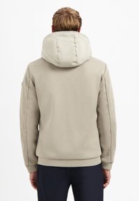 Een man die met zijn rug naar ons toe staat, draagt een beige textuurjasje met een grote gewatteerde capuchon en donkere broek tegen een eenvoudige witte achtergrond.