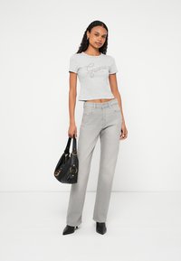 Gray cropped t-shirt with "Guess" logo, paired with light gray straight-leg jeans. Model holds a black handbag with gold accents.