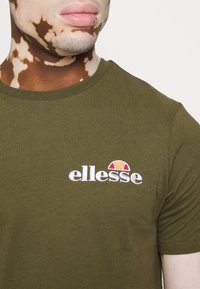 Olivegrünes T-Shirt mit kurzen Ärmeln aus Baumwolle, das das gestickte "ellesse"-Logo mit roten und orangefarbenen Akzenten über dem Text zeigt.