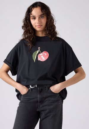 Jeune femme aux cheveux bouclés portant un t-shirt noir avec un motif cerise rouge et feuille verte, les mains dans les poches, debout devant un fond uni.