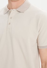 Beige polo tričko s texturovaným límcem, třemi knoflíky a prodyšnými síťovinovými prvky na rukávech, s hladkou strukturu tkaniny.