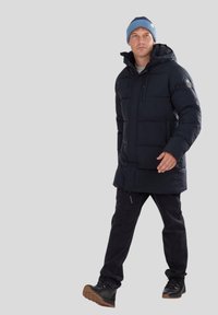 Giubbotto imbottito blu scuro con cappuccio, tessuto testurizzato, chiusura con zip, tasche laterali, abbinato a jeans neri e scarpe scure, posizionato su uno sfondo chiaro.